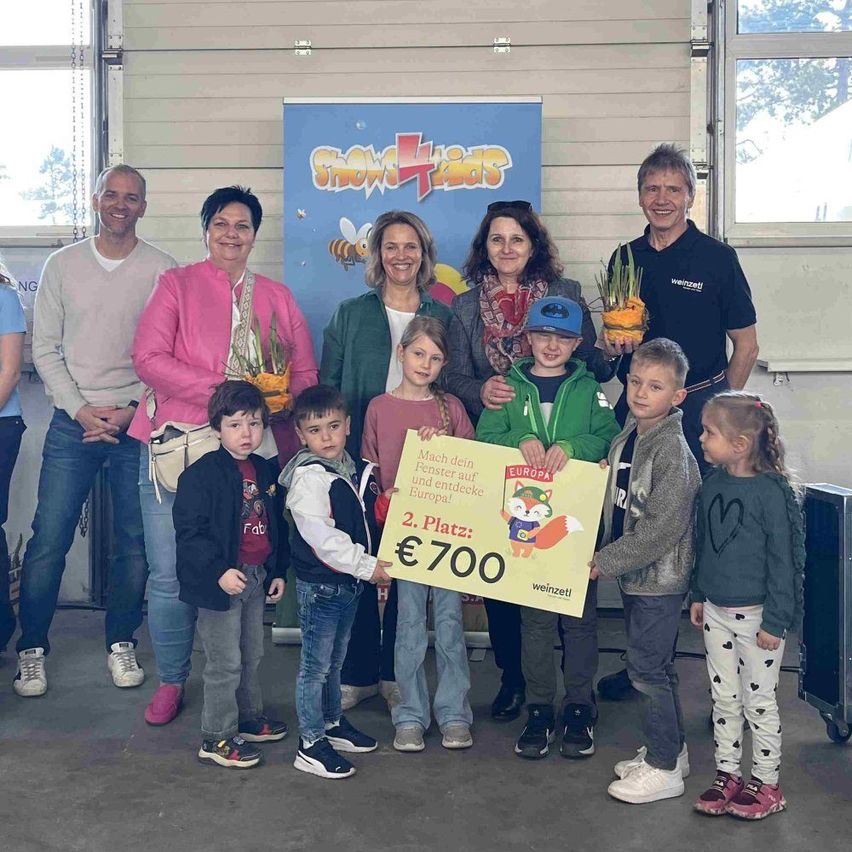 Eine Gruppe von Erwachsenen und Kindern posiert für ein Foto mit Pflanzen und einem Schild, das '2. Platz' und '€700' zeigt. Sie stehen in einem Raum mit einem blauen Banner hinter ihnen.