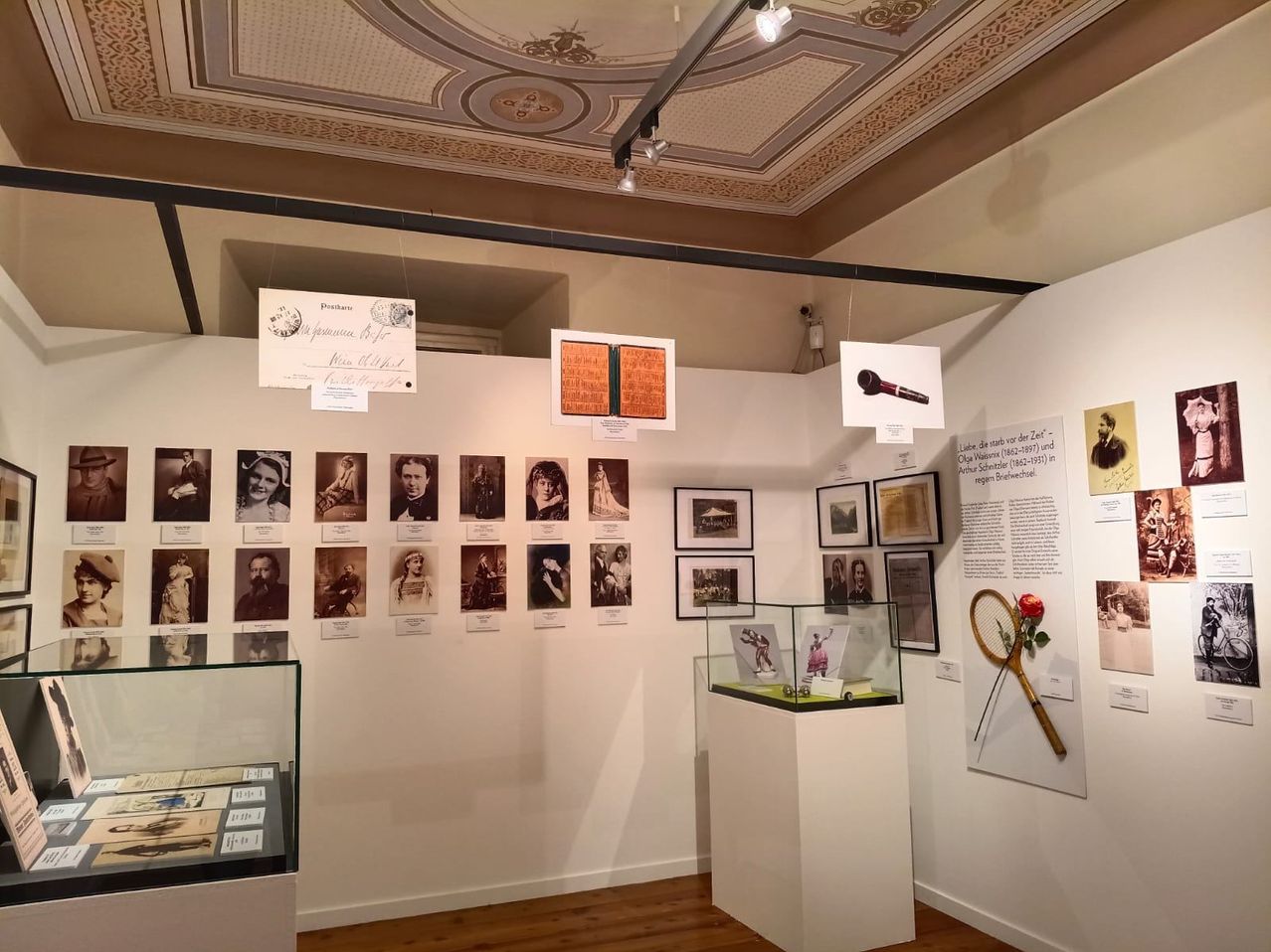 A museum exhibit displays various historical photographs, documents, and artifacts on a white wall, with a glass case in the center and decorative items hanging above.