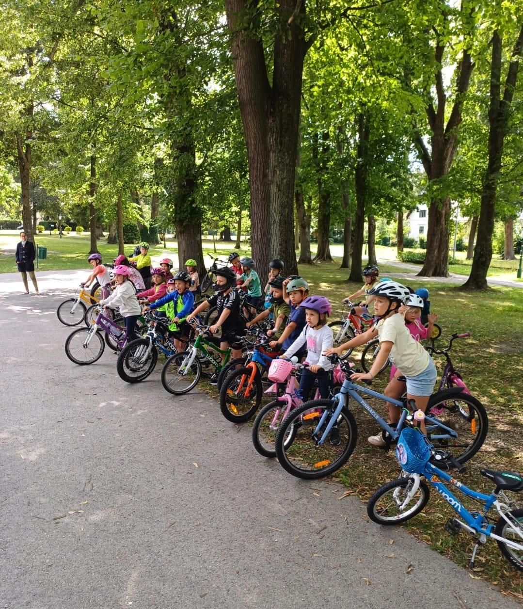 Eine Gruppe von Kindern in Helmen und Fahrradausrüstung steht mit ihren Fahrrädern in einem Park. Die Kinder sind entlang der Straßenseite aufgereiht.