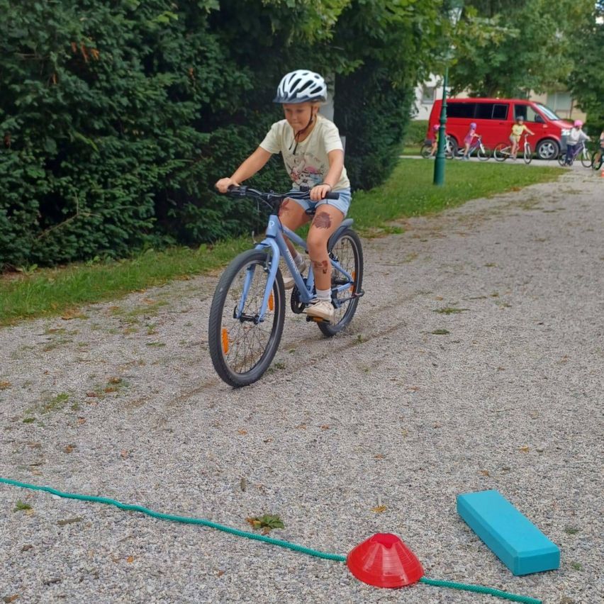 Ein junges Mädchen fährt auf einem Kiesweg ein blaues Fahrrad und trägt einen Helm. In der Nähe befinden sich rote Kegel und ein blauer Block. Im Hintergrund fahren andere Kinder Fahrrad, und ein roter Van ist geparkt.