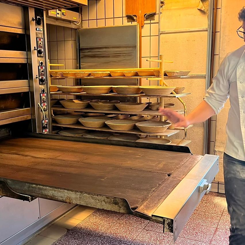 Ein Koch in einer Küche zieht einen Holztablett aus einem Ofen, hinter ihm befinden sich mehrere Regale mit Brot.