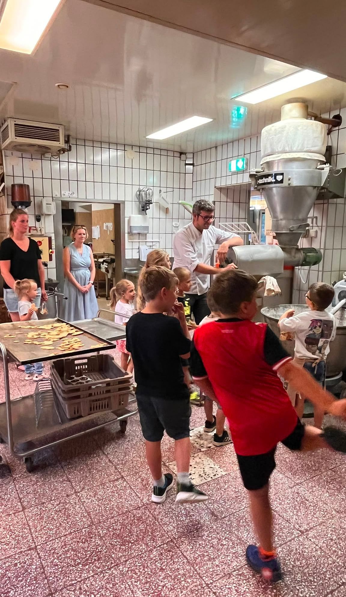 Eine Gruppe von Kindern und Erwachsenen versammelt sich in einer Küche und beobachtet einen Mann, der eine große Maschine bedient. In der Nähe stehen Tabletts mit Essen auf einem Wagen.