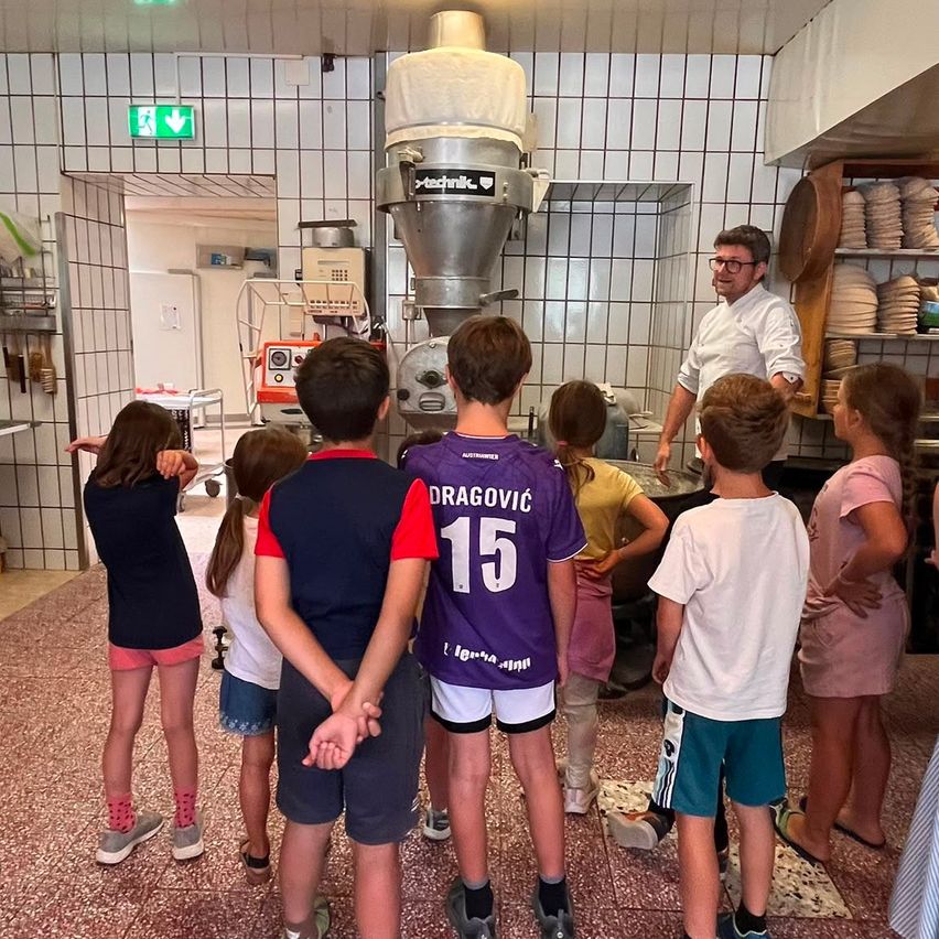 Eine Gruppe von Kindern und einem Mann sind in einer Küche. Der Mann steht vor einer großen Maschine. Die Kinder schauen sich die Maschine an.