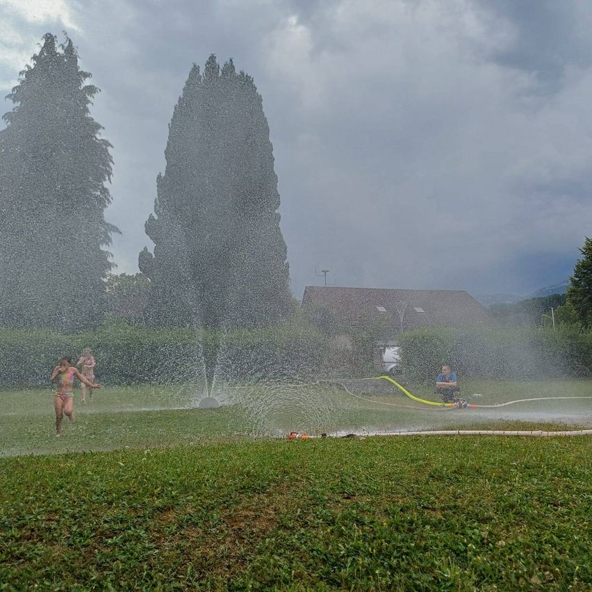 Ein Mädchen läuft durch einen Wassersprüher auf einer Wiese mit Bäumen und einem Haus im Hintergrund.
