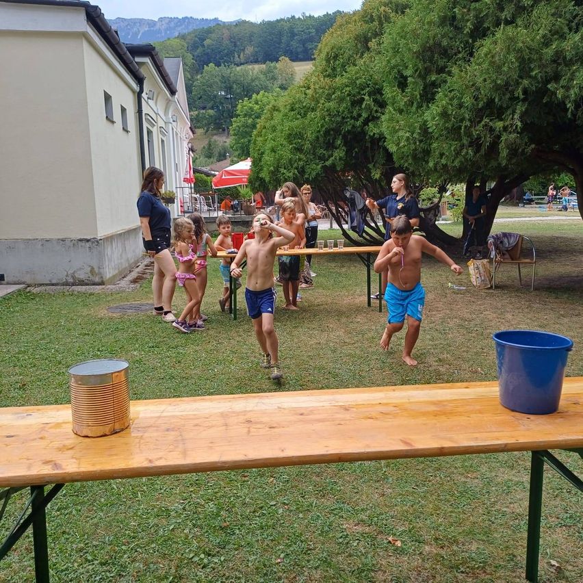 Kinder spielen ein Spiel auf einer Rasenfläche, mit einem Holztisch und Eimern in der Nähe. Eine Frau steht in der Nähe eines Gebäudes, und andere sind unter einem Baum.