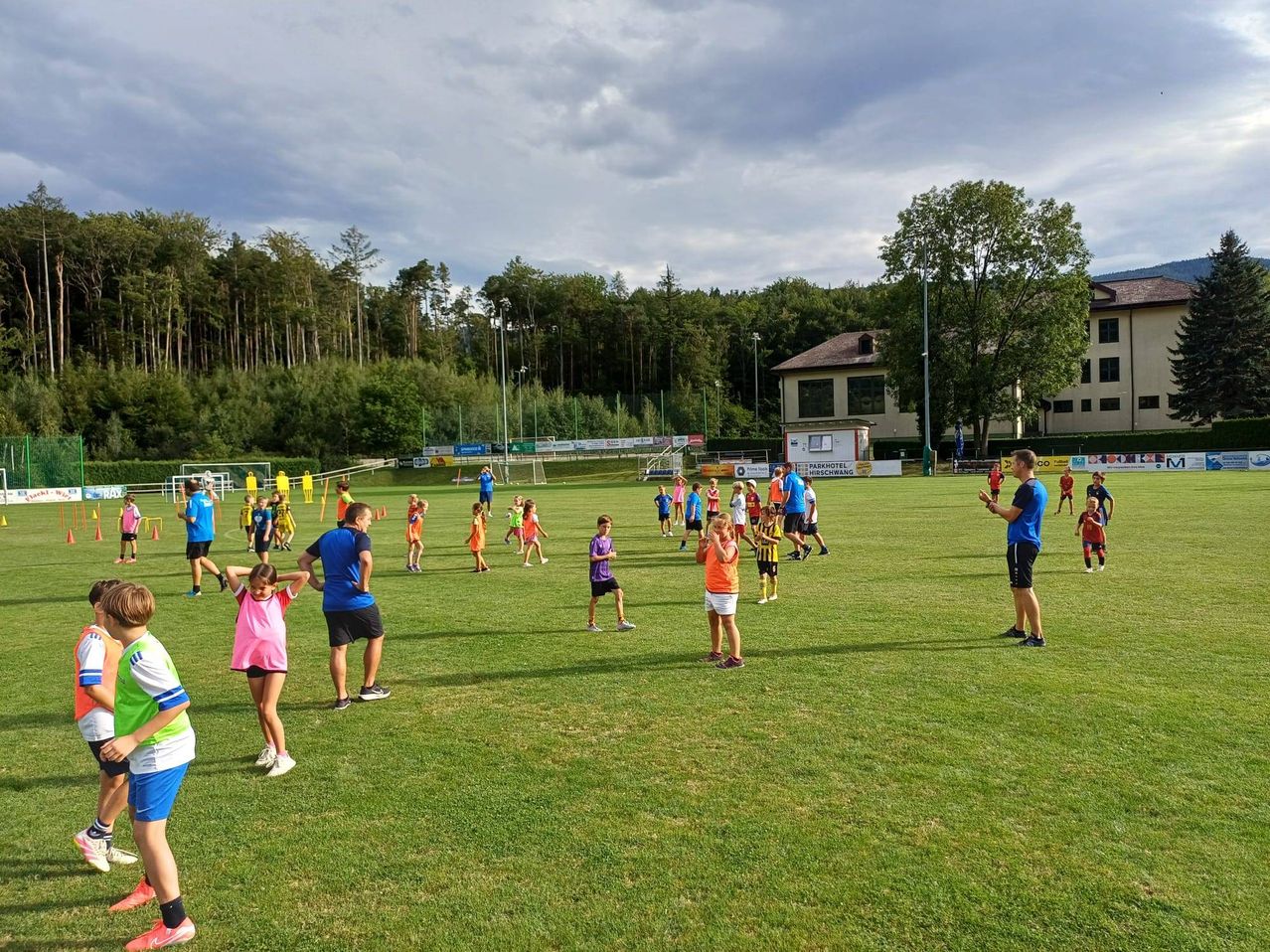 Eine Gruppe von Kindern und Erwachsenen spielt auf einem Rasenfeld bei bewölktem Himmel. Einige laufen, andere stehen. In der Ferne befinden sich Bäume und ein Gebäude.