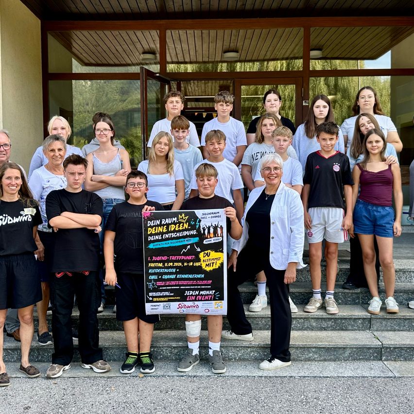 A group of students and teachers pose for a photo in front of a building. They are holding a poster with the text 'Deine Ideen Deine Entscheidung'. The students are wearing casual clothing, and some are wearing glasses. The teacher is wearing a white coat.