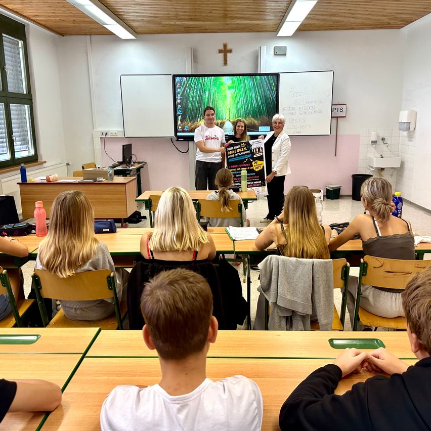 A classroom with students seated at desks, two adults standing in front with a poster, a monitor displaying an image, and a whiteboard behind them.