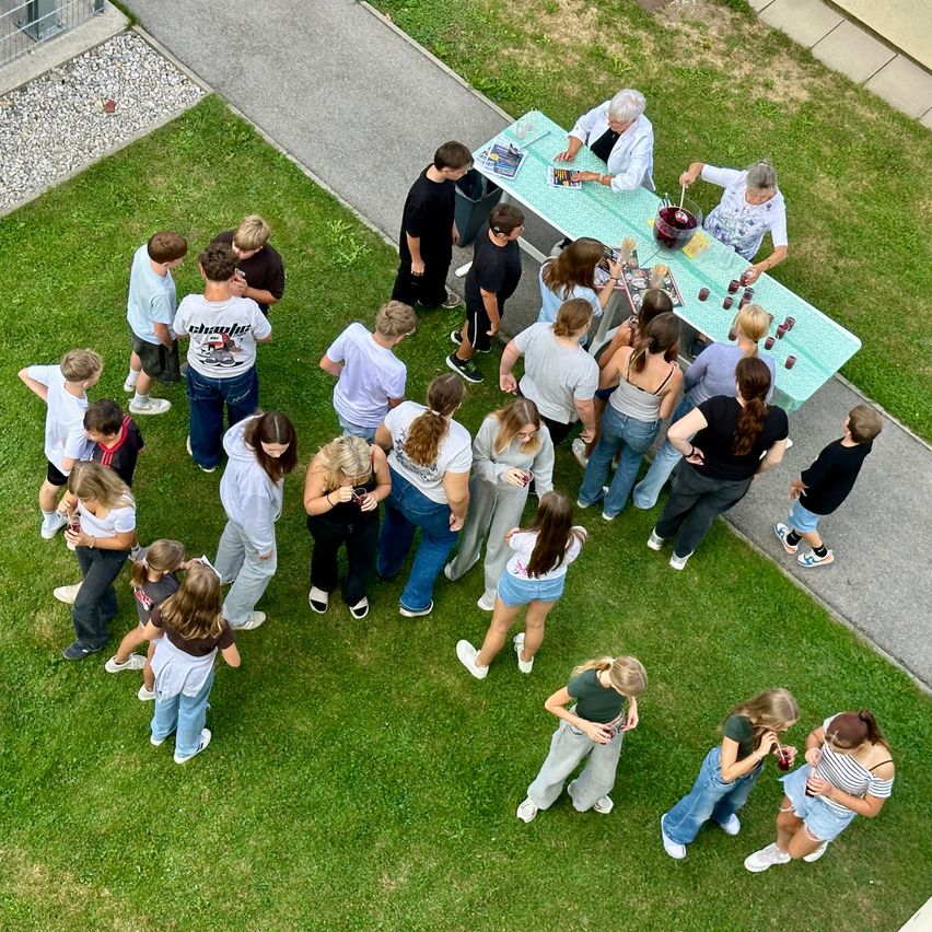 A group of people are gathered around a table with a red bowl, possibly preparing food or drinks. Two older women are standing at the table. The group is on a grassy area with a pathway nearby.