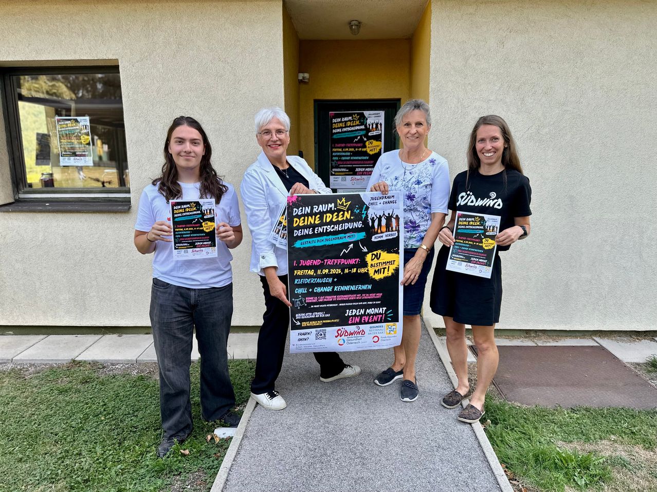 Four women stand in front of a building, holding a large poster with colorful text and images. They are smiling and appear to be posing for a photo.