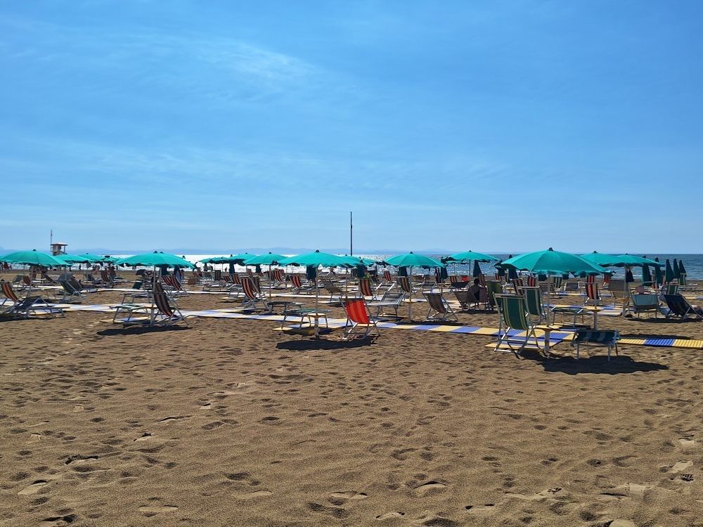 Ein sonniger Strand mit vielen Menschen, die sich unter türkisfarbenen Sonnenschirmen entspannen. Der Sand ist golden und der Himmel klar.