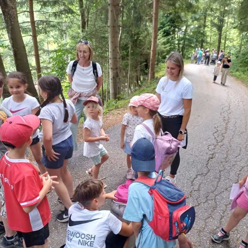A group of children and adults are gathered on a path in a forest, with two women standing at the front. The children are wearing hats and backpacks, and some are holding papers. The women are smiling and appear to be leading a group activity.