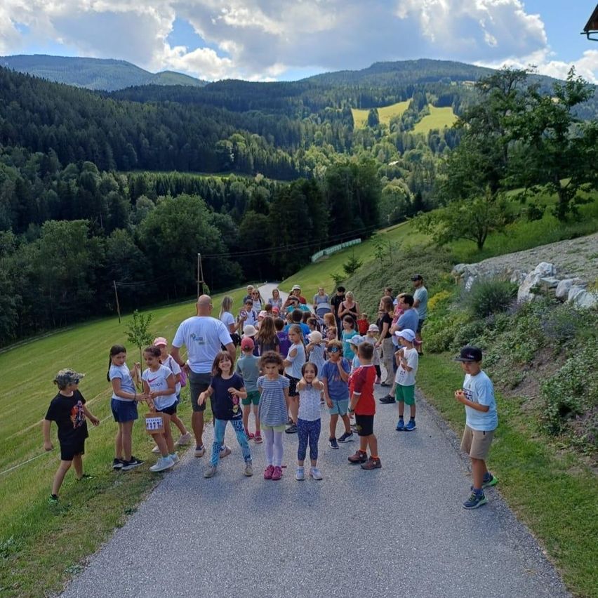 A group of children and adults stand on a pathway in a grassy field with mountains in the background. Some are wearing hats and sneakers, and a man holds a small tree.