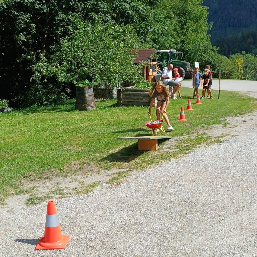A young girl pushes a wheelbarrow filled with colorful balls down a path lined with traffic cones. In the background, several people are standing near a tractor and a wooden structure.