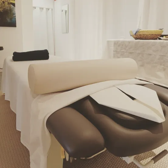 Ein weißer Stoff bedeckt einen braunen Massagestuhl. Ein aufgerolltes weißes Handtuch ruht darauf, mit einem schwarzen Handtuch auf der linken Seite. Ein Spiegel und ein geflochtener Korb sind im Hintergrund.