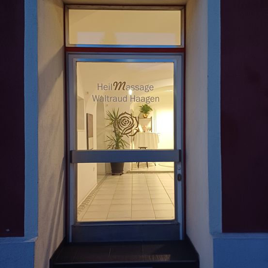 Die Glastür eines Massagestudios steht offen. Darauf steht 'Heilmassage Waltraud Haagen'. Eine Topfpflanze steht in der Ecke des Raumes.