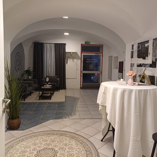 Ein Flur mit einem gewölbten Dach führt zu einem Raum mit Sofas, einem Tisch und einem Teppich. Eine Tür ist offen und zeigt einen hellen Raum. Ein Tisch mit weißem Tuch und Blumenvase steht im Vordergrund.