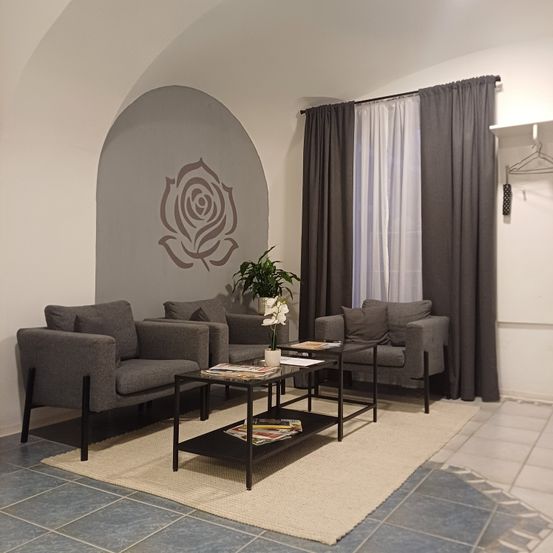 Ein Wohnzimmer mit zwei grauen Sesseln und einem Couchtisch auf einem beigen Teppich. Eine Wand mit Rosendesign und graue Vorhänge schmücken den Raum. Ein Topf mit Pflanze steht auf dem Tisch.