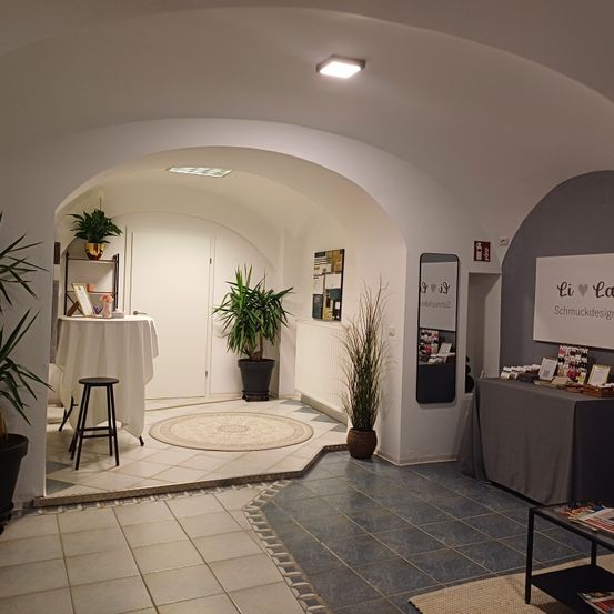 Ein moderner Innenbereich mit weißen Wänden und gewölbten Eingängen, ausgestattet mit einem Tisch mit weißer Tischdecke, einem Hocker und Topfpflanzen. Rechts zeigt eine Wand 'Li La Schmuckdesign', mit zusätzlichen Artikeln auf einem Tisch.