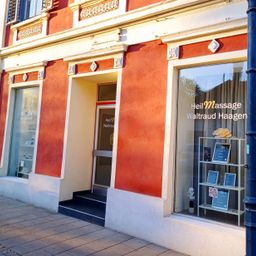 Ein rotes Gebäude mit einem Glasfenster und einer Tür, auf der 'Heil Massage Waltraud Haagen' steht. Ein kleiner Topf mit einer Pflanze und eine gelbe Rose sind im Fenster.