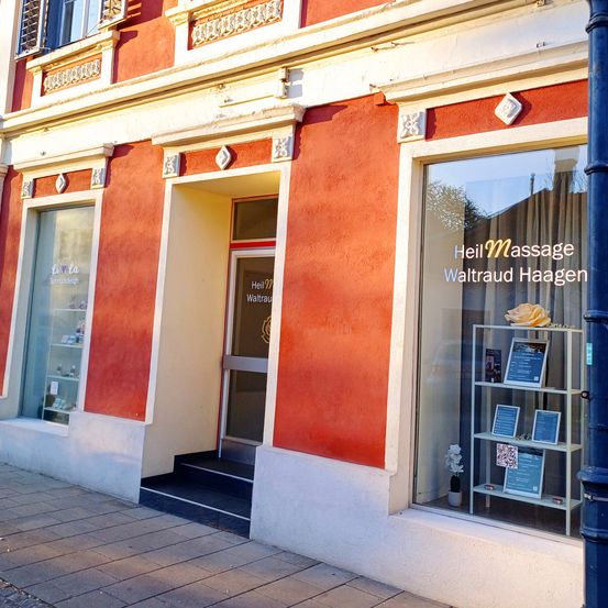 Ein rotes Gebäude mit einem Glasfenster und einer Tür, auf der 'Heil Massage Waltraud Haagen' steht. Ein kleiner Topf mit einer Pflanze und eine gelbe Rose sind im Fenster.