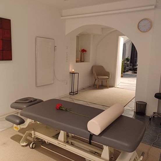 Ein sauberer, moderner Therapieraum mit einem Massagetisch und einer roten Rose, weißen Wänden und einer Uhr an der Wand.