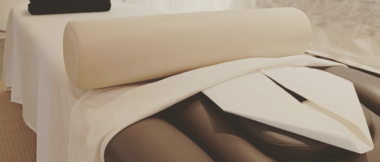 Eine Person liegt auf einem Bett mit einem aufgerollten beigen Kissen über den Beinen, bedeckt von einem weißen Tuch. Ein schwarzes Objekt befindet sich oben links.