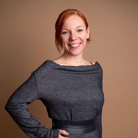 Eine Frau mit roten Haaren lächelt und posiert für ein Foto. Sie trägt ein graues Kleid mit einem schwarzen Gürtel und hat eine Hand auf der Hüfte.