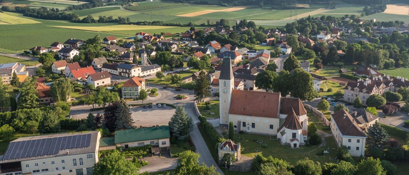 Luftaufnahme eines ländlichen Dorfes mit einer Kirche, Wohnhäusern und grünen Feldern. Das Dorf ist von üppigen Bäumen und Bergen umgeben.