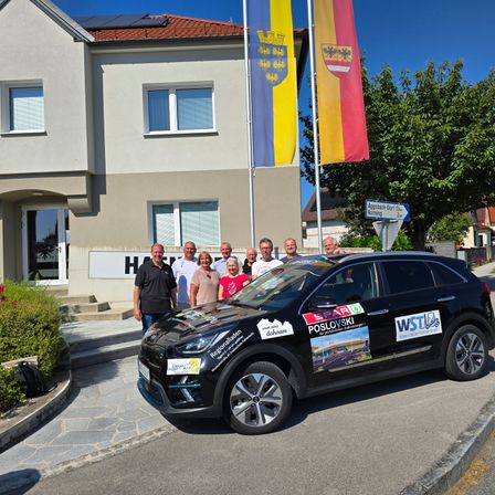 Eine Gruppe von Menschen steht vor einem Gebäude mit Flaggen und einem Auto mit verschiedenen Aufklebern. Das Gebäude hat Solarmodule auf dem Dach und befindet sich an einer Straße.