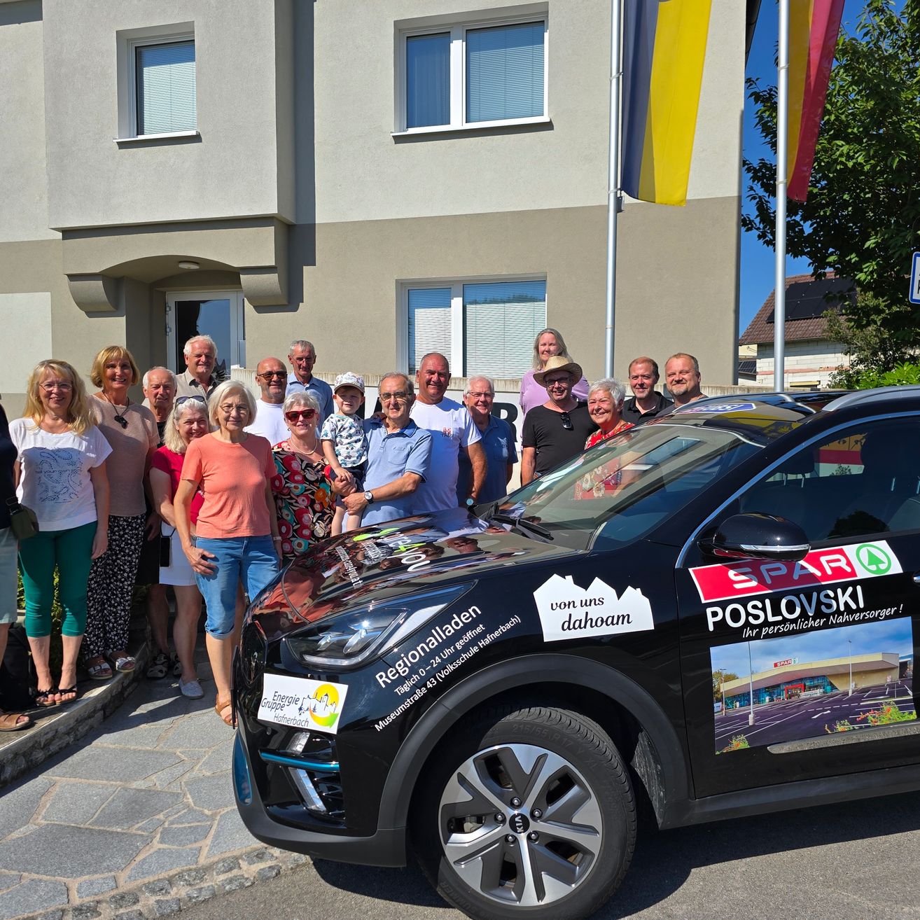 Eine Gruppe von Menschen steht vor einem modernen Gebäude und posiert für ein Foto in der Nähe eines schwarzen Autos mit regionalen Werbeaufklebern. Das Auto ist auf der Straße geparkt, mit Fahnen dahinter.