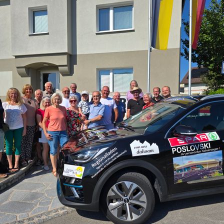 Eine Gruppe von Menschen steht vor einem modernen Gebäude und posiert für ein Foto in der Nähe eines schwarzen Autos mit regionalen Werbeaufklebern. Das Auto ist auf der Straße geparkt, mit Fahnen dahinter.