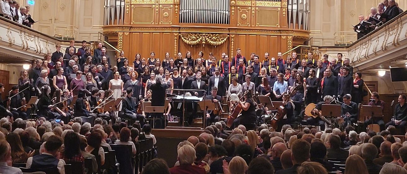 Ein großes Orchester spielt auf einer Bühne vor einer großen Orgel mit Kronleuchtern darüber, das Publikum sitzt unten.
