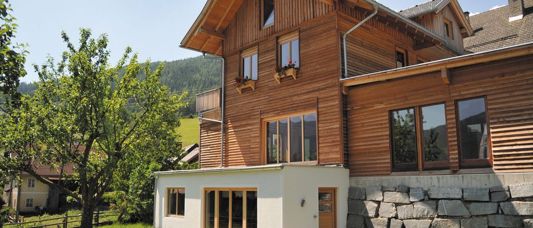 Ein modernes Holzhaus mit Steinmauerfundament, Glasfenstern und zwei Balkonen mit Blumenkästen an einem sonnigen Tag.