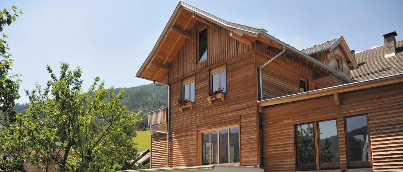 Außenansicht eines modernen Holzhauses mit großen Fenstern, umgeben von Grün, Bergen und einem klaren blauen Himmel.