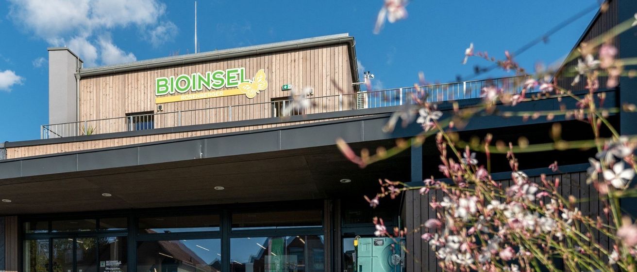Die Vorderseite eines Bioinsel-Ladens mit einem gelben Schmetterlingslogo, Einkaufswagen und einem Mülleimer draußen an einem sonnigen Tag.