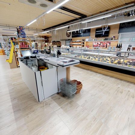 Ein Supermarkt mit einer Kasse vor einer Vitrine voller Käse, einem Regal mit Produkten und einem Monitor.
