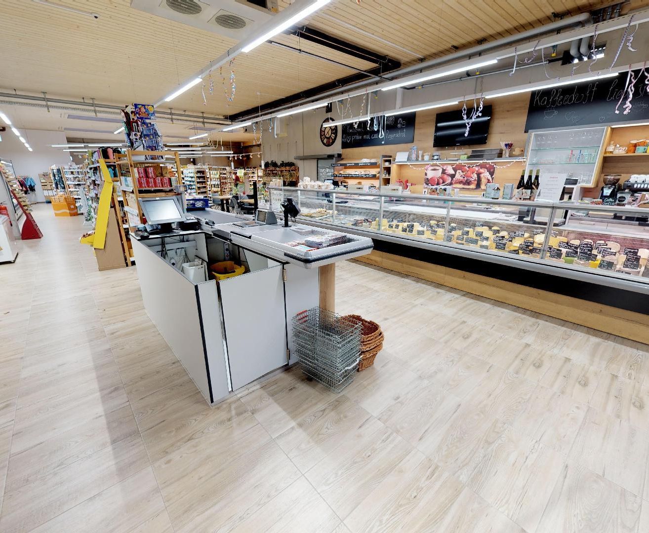 Ein Supermarkt mit einer Kasse vor einer Vitrine voller Käse, einem Regal mit Produkten und einem Monitor.
