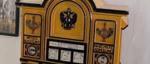 Ein gelber Postkasten ist an der Wand montiert. Er hat eine Uhr und den Text 'nachste Abholung' auf der Vorderseite. Darüber ist eine Karte und ein Bilderrahmen montiert.