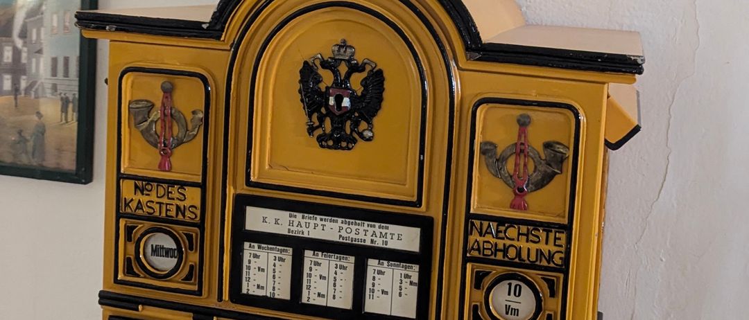 Ein gelber Postkasten ist an der Wand montiert. Er hat eine Uhr und den Text 'nachste Abholung' auf der Vorderseite. Darüber ist eine Karte und ein Bilderrahmen montiert.