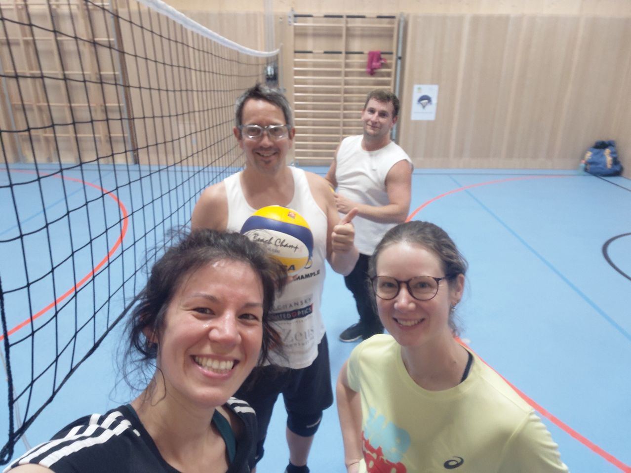 Four people stand in a gym, smiling. One man holds a volleyball. Behind him, two other men stand, one smiling and giving a thumbs up. The floor is blue, and wooden walls are visible.