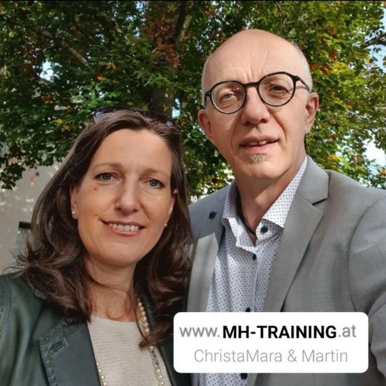 Ein Mann und eine Frau stehen zusammen draußen vor einem Baum. Der Mann trägt eine Brille und einen grauen Anzug, und die Frau trägt eine Halskette und eine schwarze Jacke. Es gibt einen Wasserzeichen mit dem Text www.MH-TRAINING.at ChristaMara & Martin.