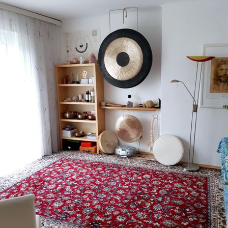 Ein ruhiger Raum mit einem großen Gong an der Wand. Ein Holzregal mit verschiedenen Gegenständen, einschließlich Schüsseln, steht in der Nähe. Ein roter Teppich bedeckt den Boden, und eine Lampe steht rechts.