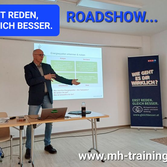 Ein Mann steht vor einem Tisch und hält eine Präsentation. Eine Projektionsleinwand zeigt eine Folie mit der Überschrift 'Energiequellen erkennen & nutzen.' Ein Laptop, eine Flasche und Papiere liegen auf dem Tisch. Ein Banner rechts sagt 'Wie geht es dir wirklich?' mit einer Website. Der Raum hat Holzböden.