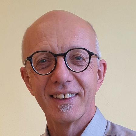 Ein älterer Mann mit kahlgeschorenem Kopf und Schnurrbart, der eine Brille und ein blaues Hemd mit Kragen trägt, lächelt fröhlich.