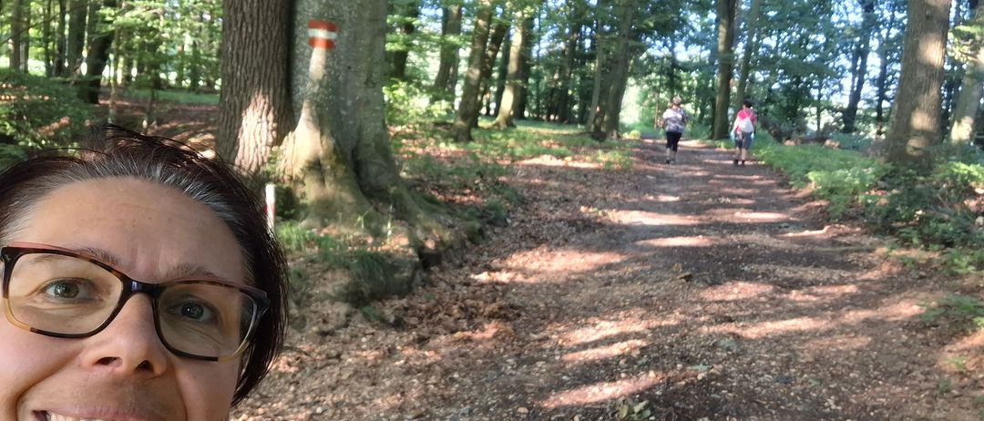 Eine Frau macht ein Selfie auf einem Waldweg, mit zwei Personen, die auf dem Pfad vor ihr gehen. Der Weg ist von hohen Bäumen umgeben, und die Sonne scheint durch die Bäume.