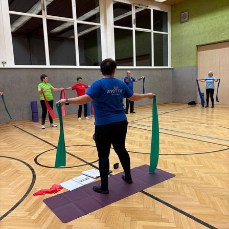Eine Gruppe Erwachsener in einer Turnhalle, die ein Training mit Widerstandsbändern durchführt. Eine Person führt, während andere im Hintergrund folgen.