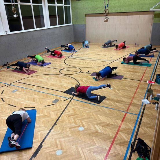 Eine Gruppe von Menschen trainiert auf einem Holzboden, einige liegen auf Matten. Andere machen Liegestütze, und es sind verschiedene Trainingsgeräte herum verstreut. Der Raum hat weiße Wände und Fenster.