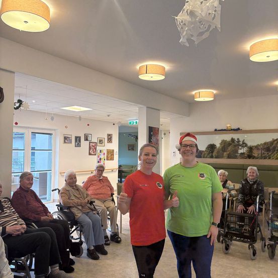 Zwei Frauen zeigen Daumen hoch in einem Raum mit älteren Menschen, die auf Stühlen und Rollstühlen sitzen. Der Raum ist mit Weihnachtsdekoration geschmückt.