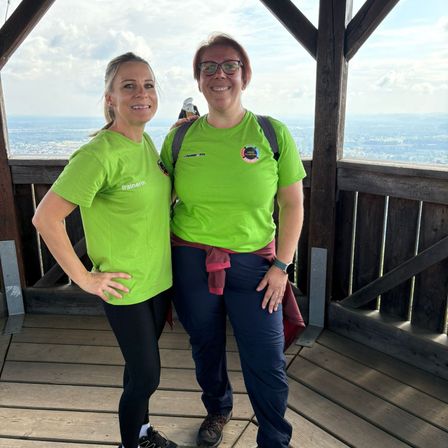 Zwei Frauen in grünen Shirts und mit Brille stehen lächelnd auf einer hölzernen Plattform zur Kamera, mit Blick auf eine Stadtlandschaft im Hintergrund.